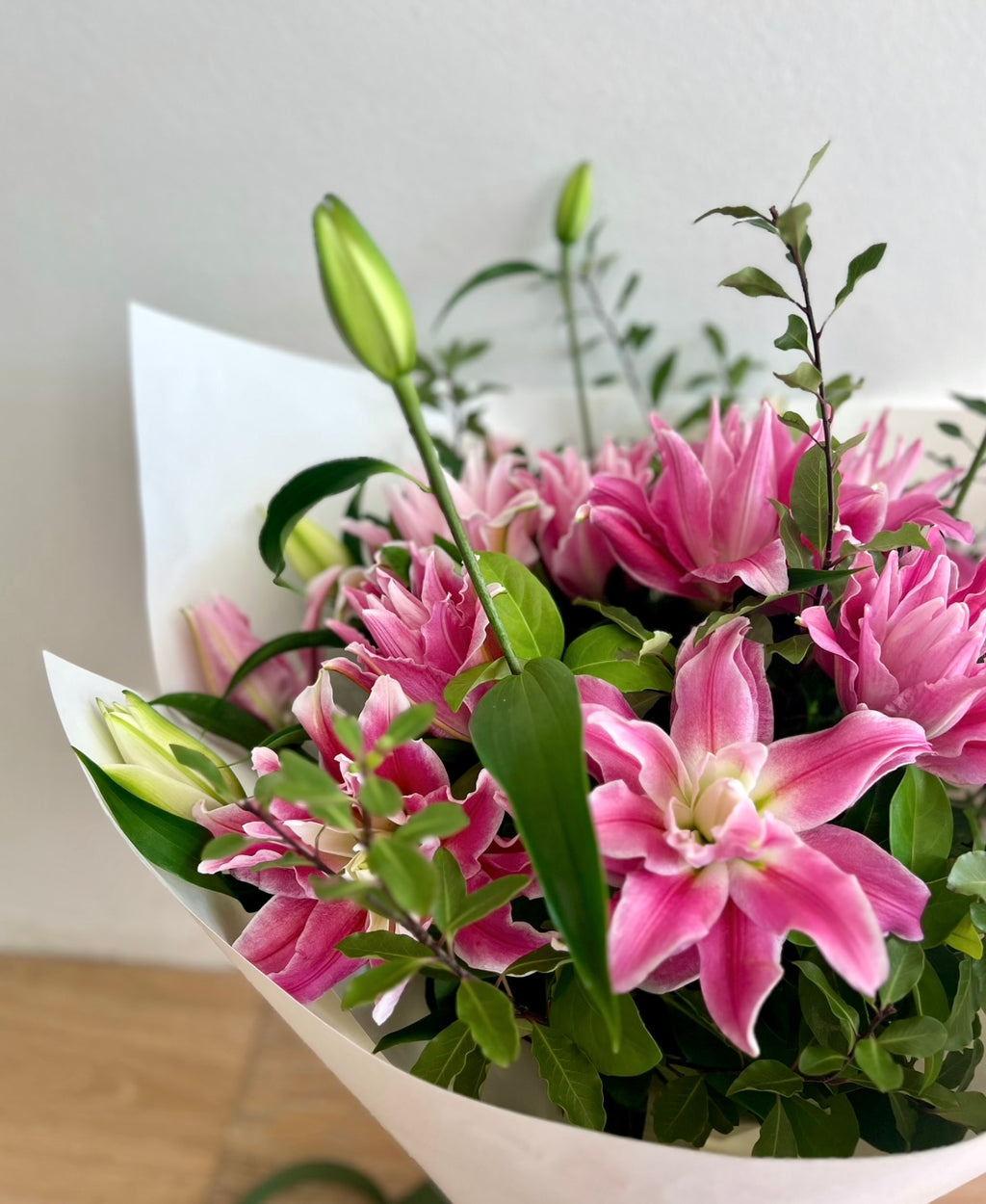 A hand-tied bouquet of pink lilies with green foliage, wrapped in white paper with a ribbon.