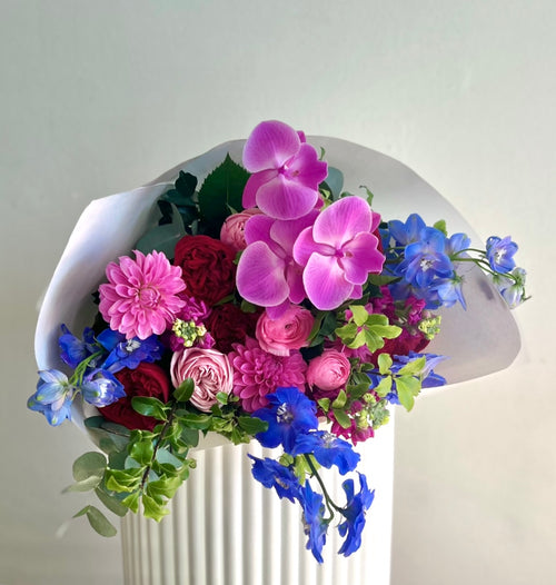 Jewel toned bouquet of flowers including orchids wrapped in white paper and a ribbon.